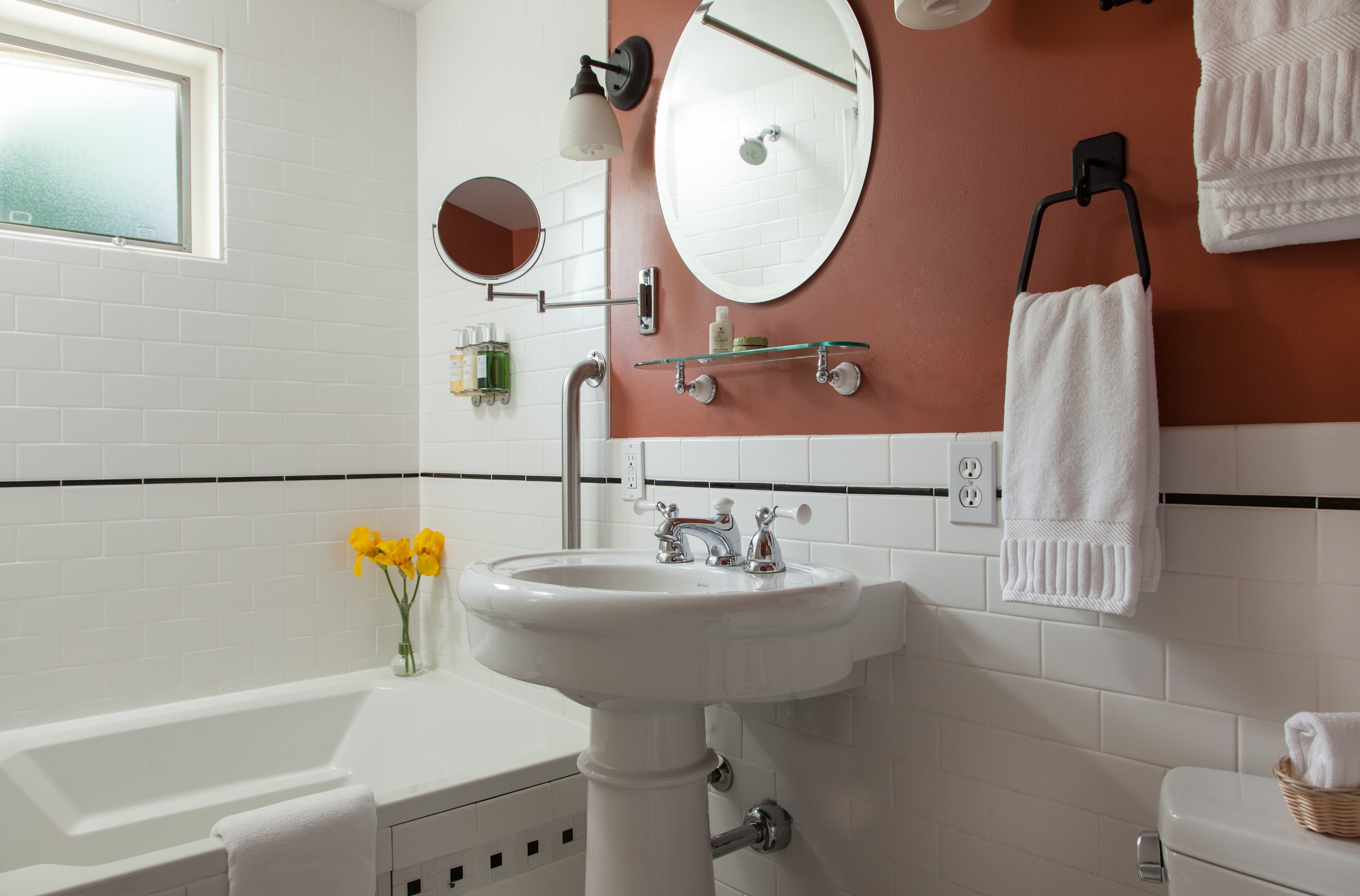 Modern bathroom with a white pedestal sink, round mirrors, terracotta wall above subway tiles, and yellow flowers.
