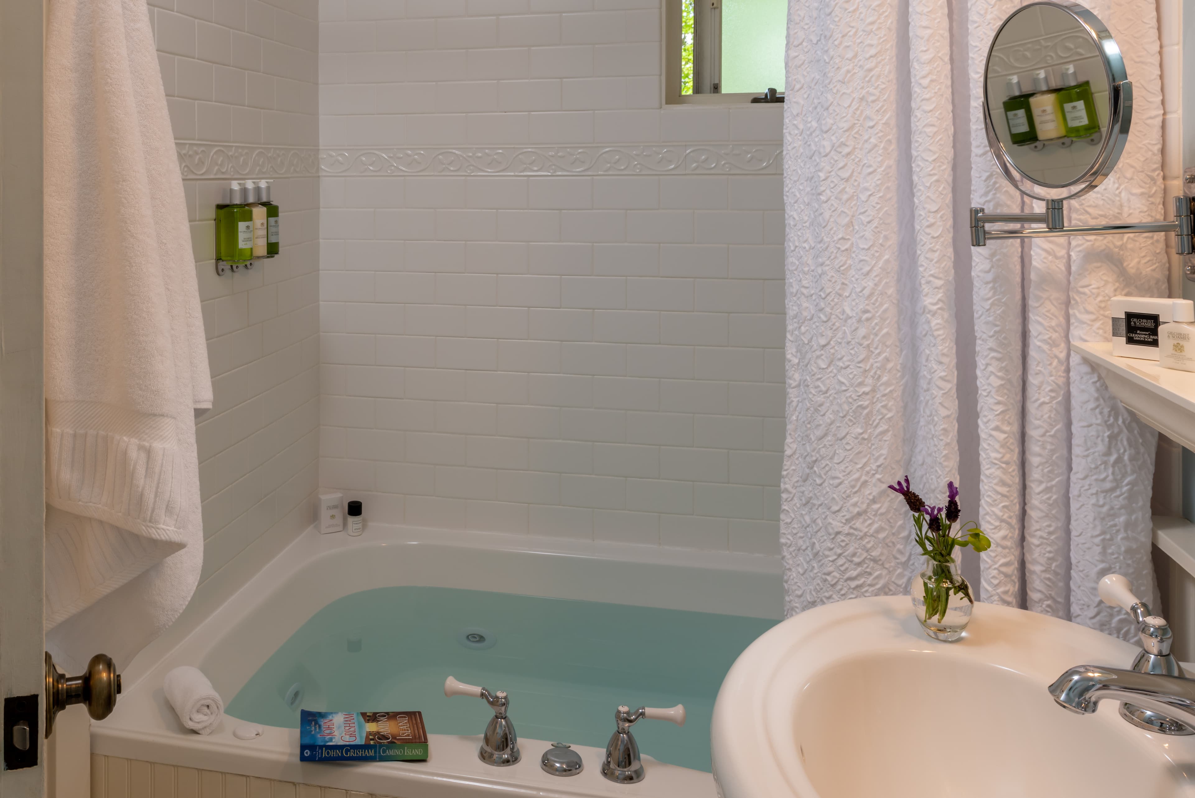 A bathroom with a white tiled wall, half-filled bathtub, hanging towel, and amenities on the ledge.