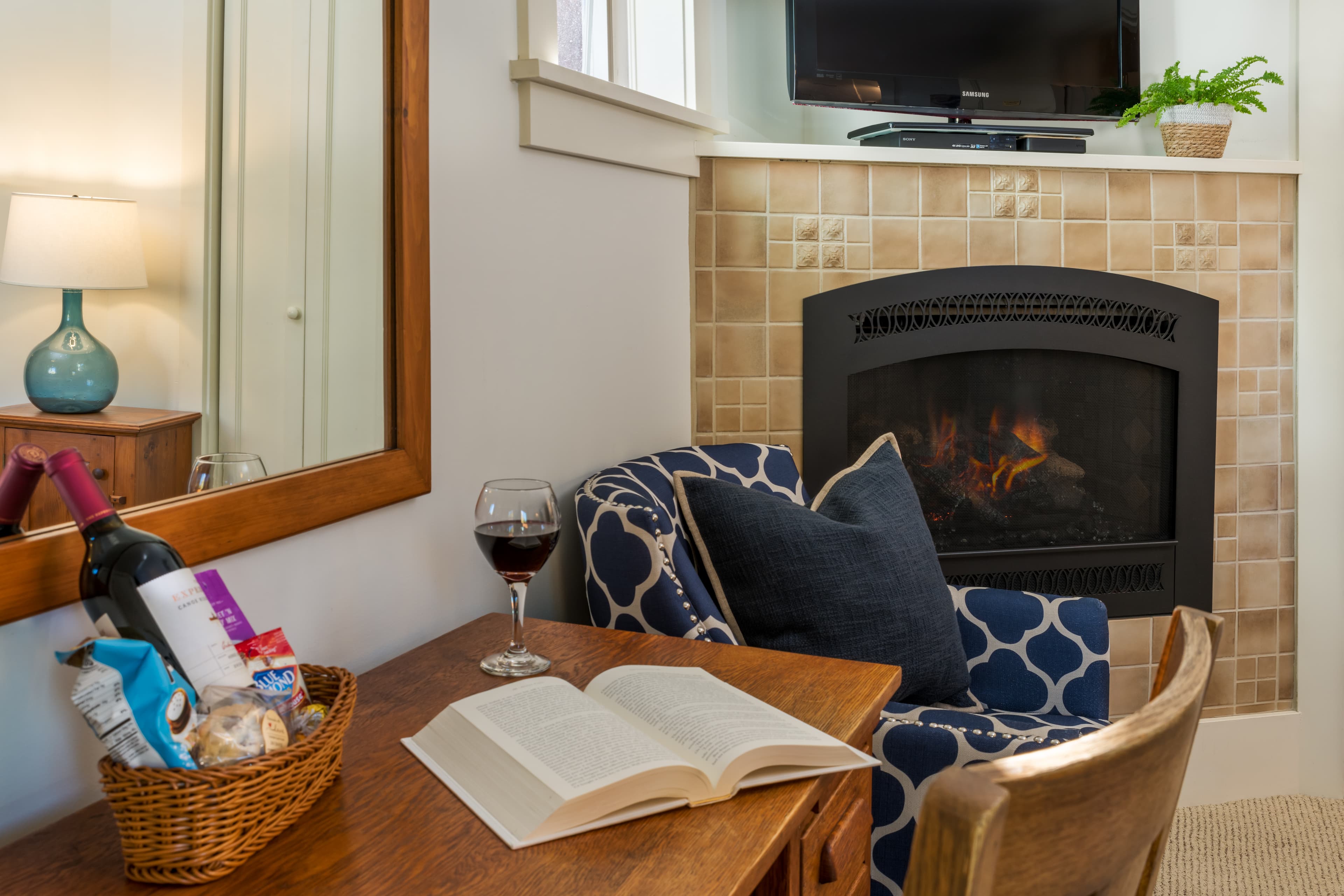 Cozy reading nook with an open book, wine glass, patterned blue chair, and lit fireplace.