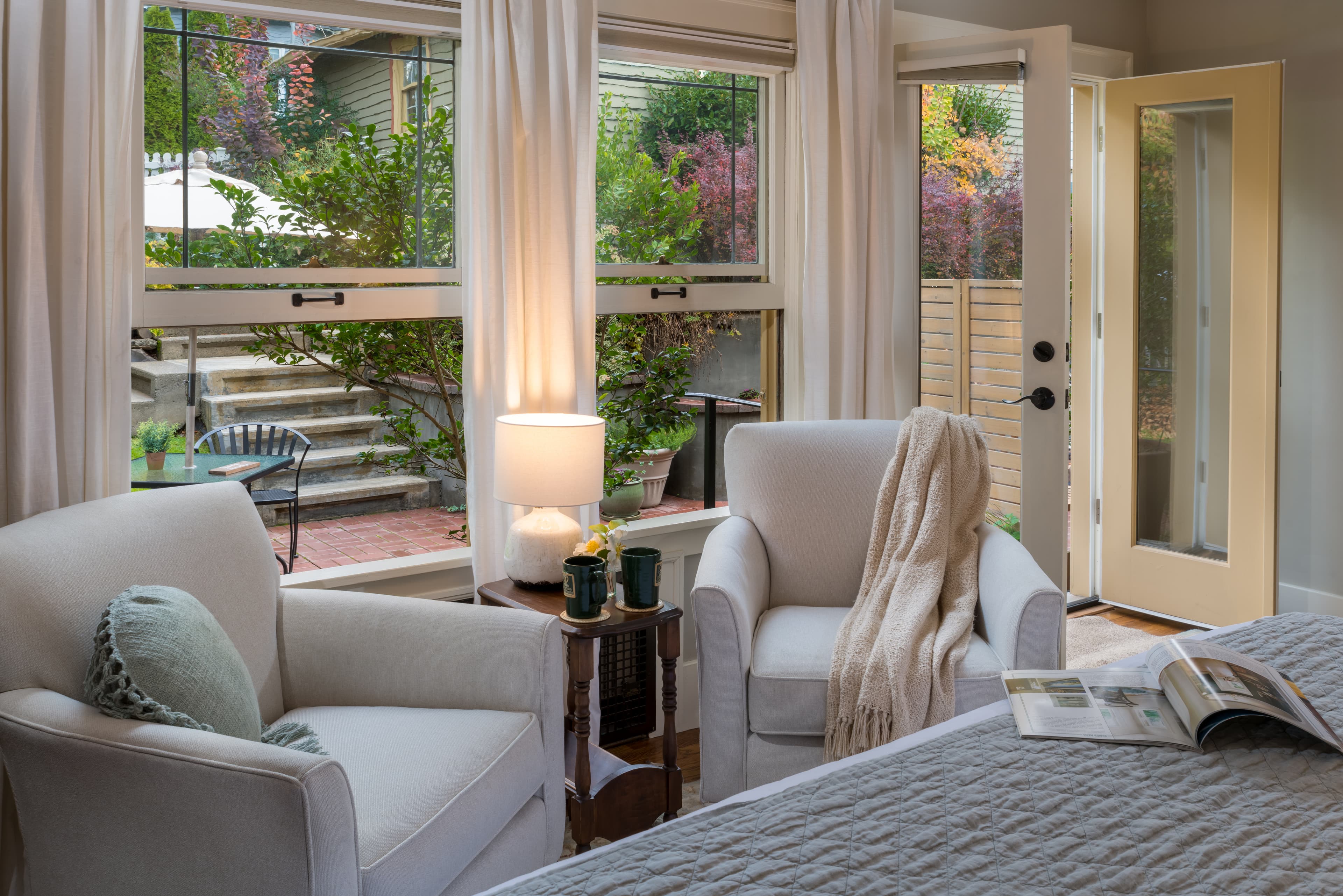 Cozy living room with two white armchairs, a lamp, and open windows overlooking greenery.