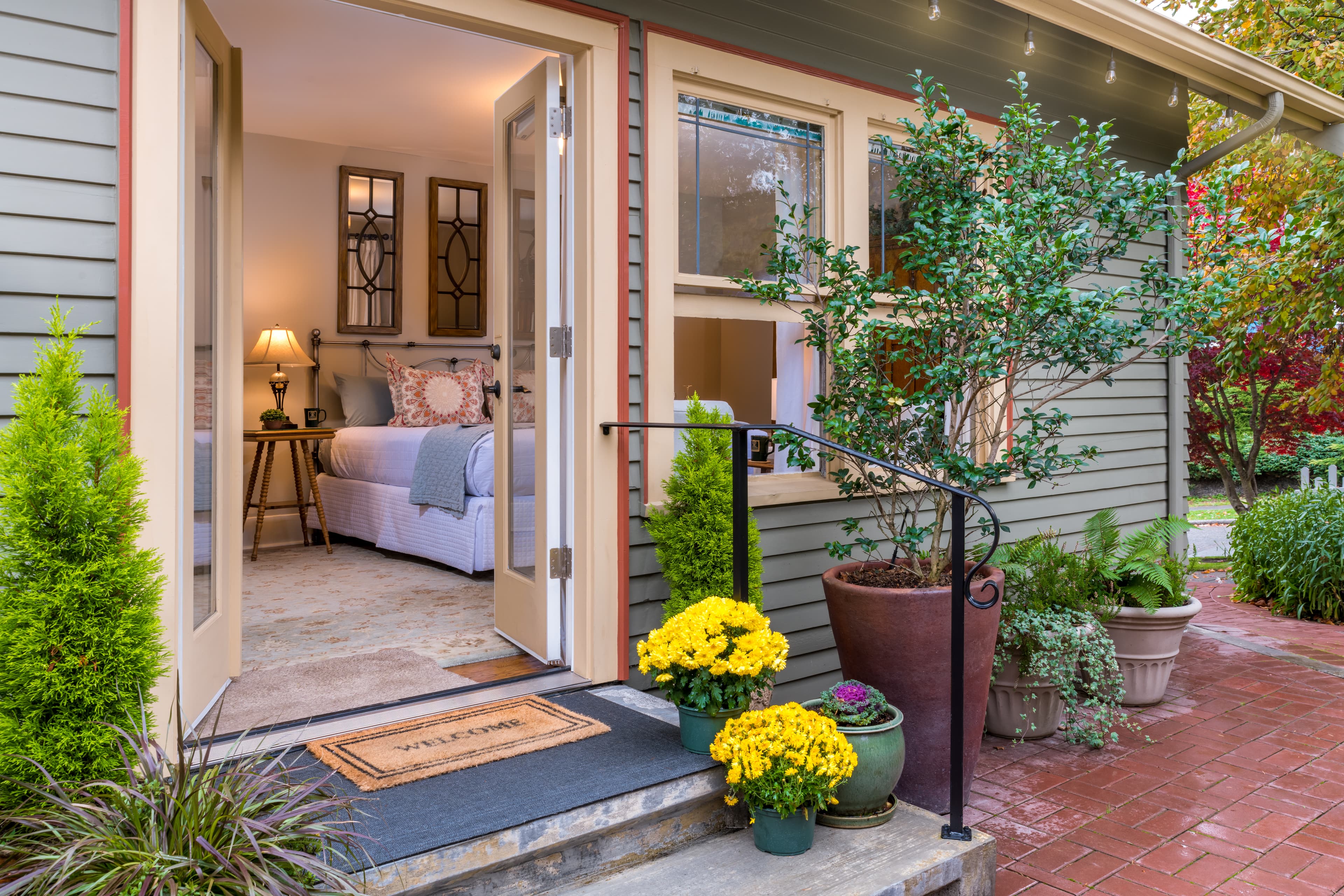Cozy bedroom visible through an open door from a welcoming porch with plants and a welcome mat.