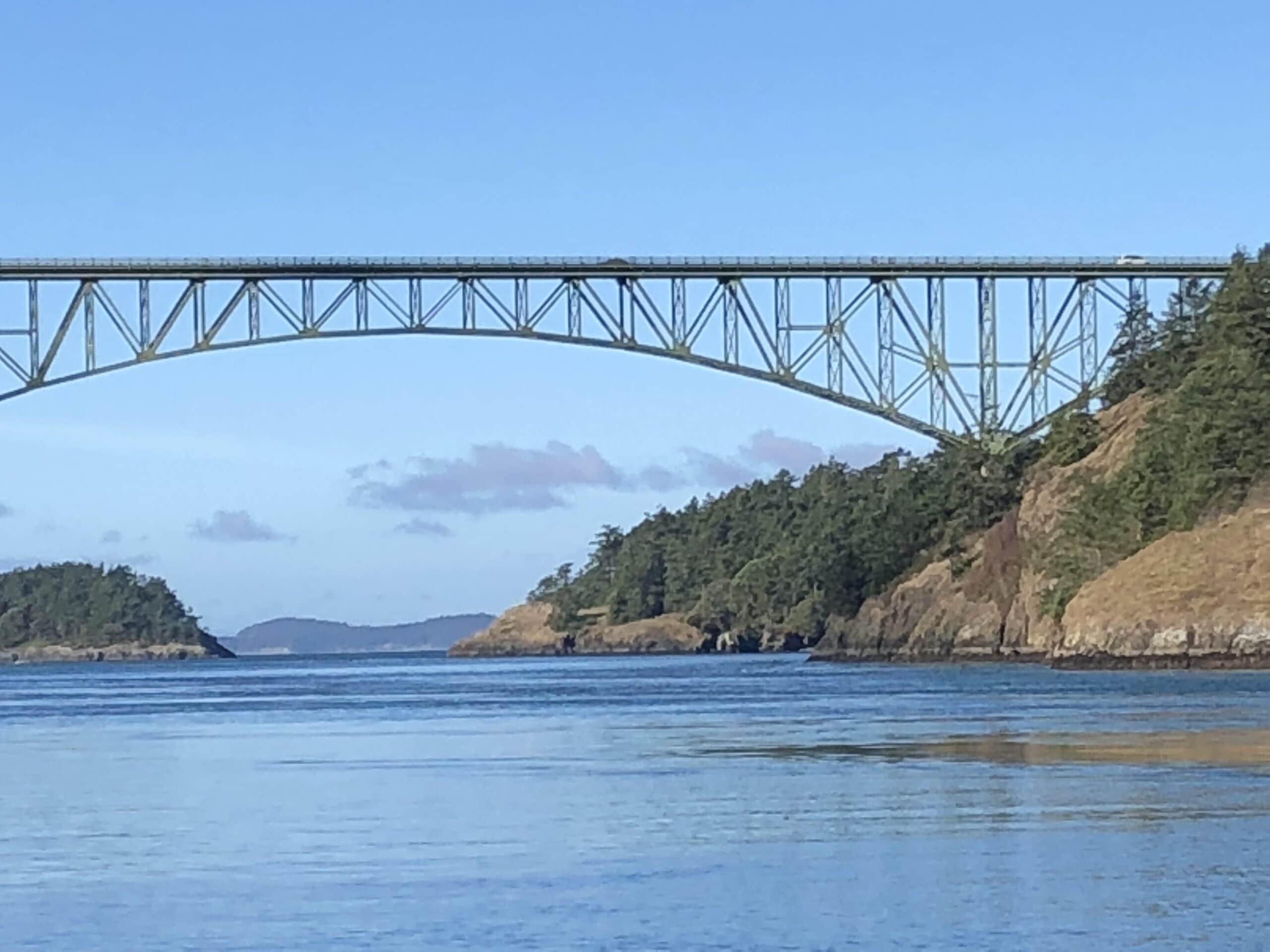 A steel bridge spans over calm water, surrounded by forested hills and distant islands under a clear blue sky.