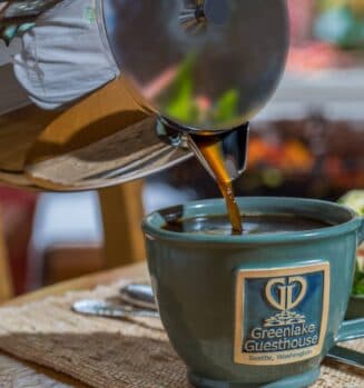 A coffee pot is pouring coffee into a blue mug with a "Greenlake Guesthouse" logo.