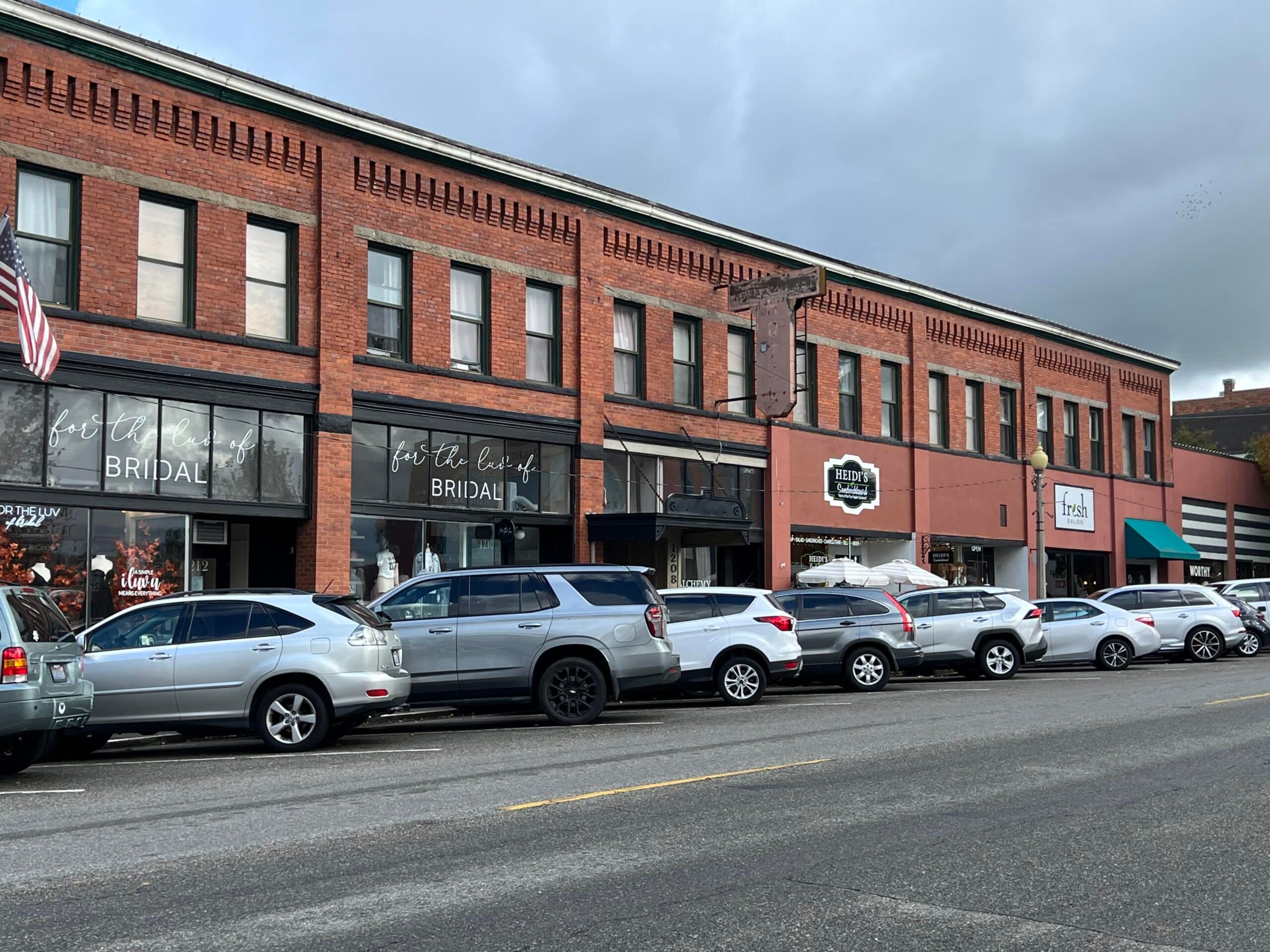 A row of brick storefronts with various businesses and parked cars along the street.