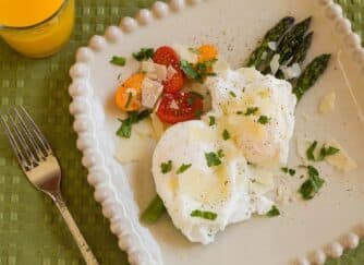 Plate of two poached eggs on asparagus, accompanied by cherry tomatoes and garnished with herbs, with a glass of orange juice in the background.