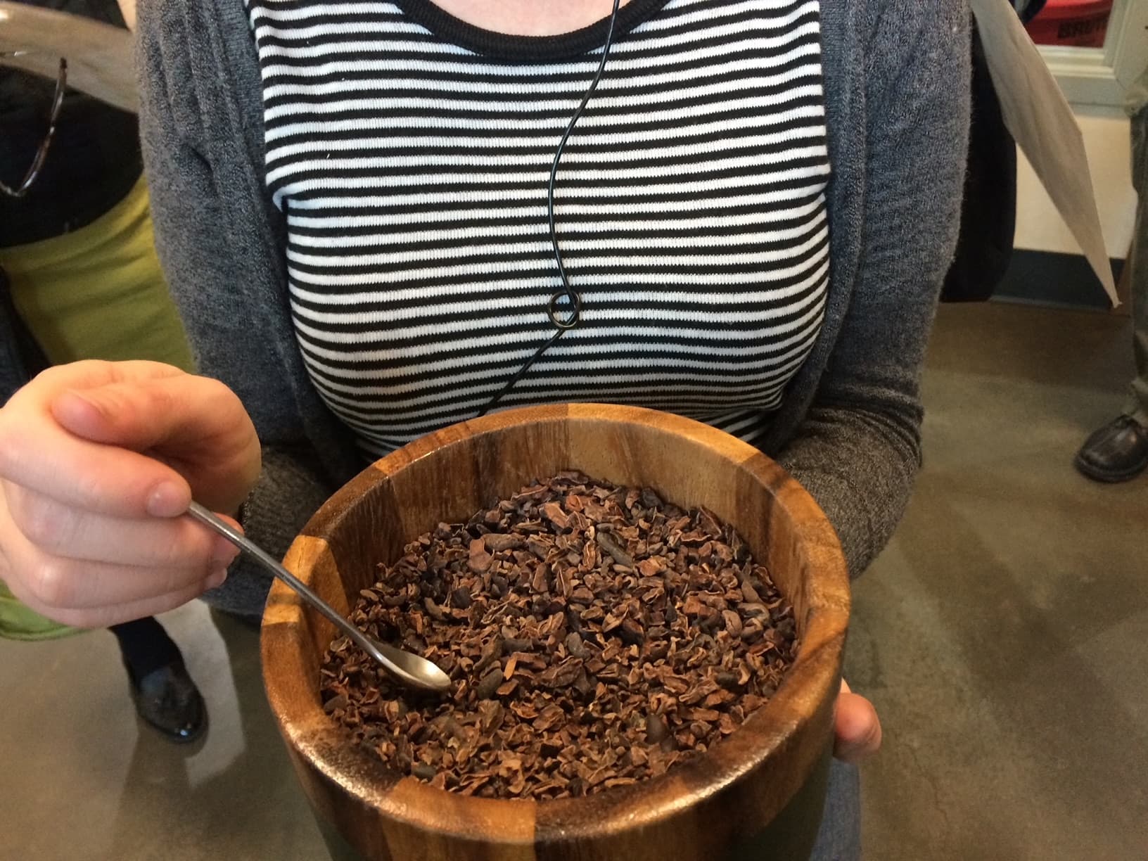 A person holds a wooden bowl filled with cocoa nibs, with a spoon resting inside.