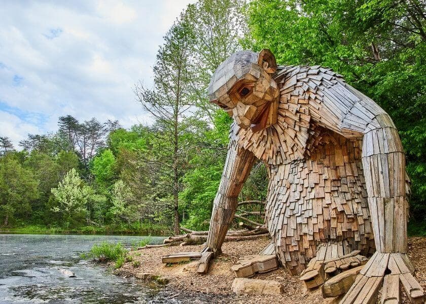 A large wooden sculpture of a troll-like figure sits by a pond surrounded by trees.