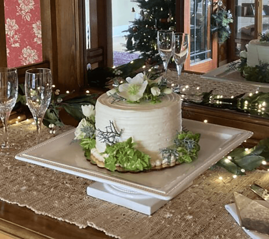 A decorated cake with greenery and flowers on a table, surrounded by champagne glasses and holiday decor.