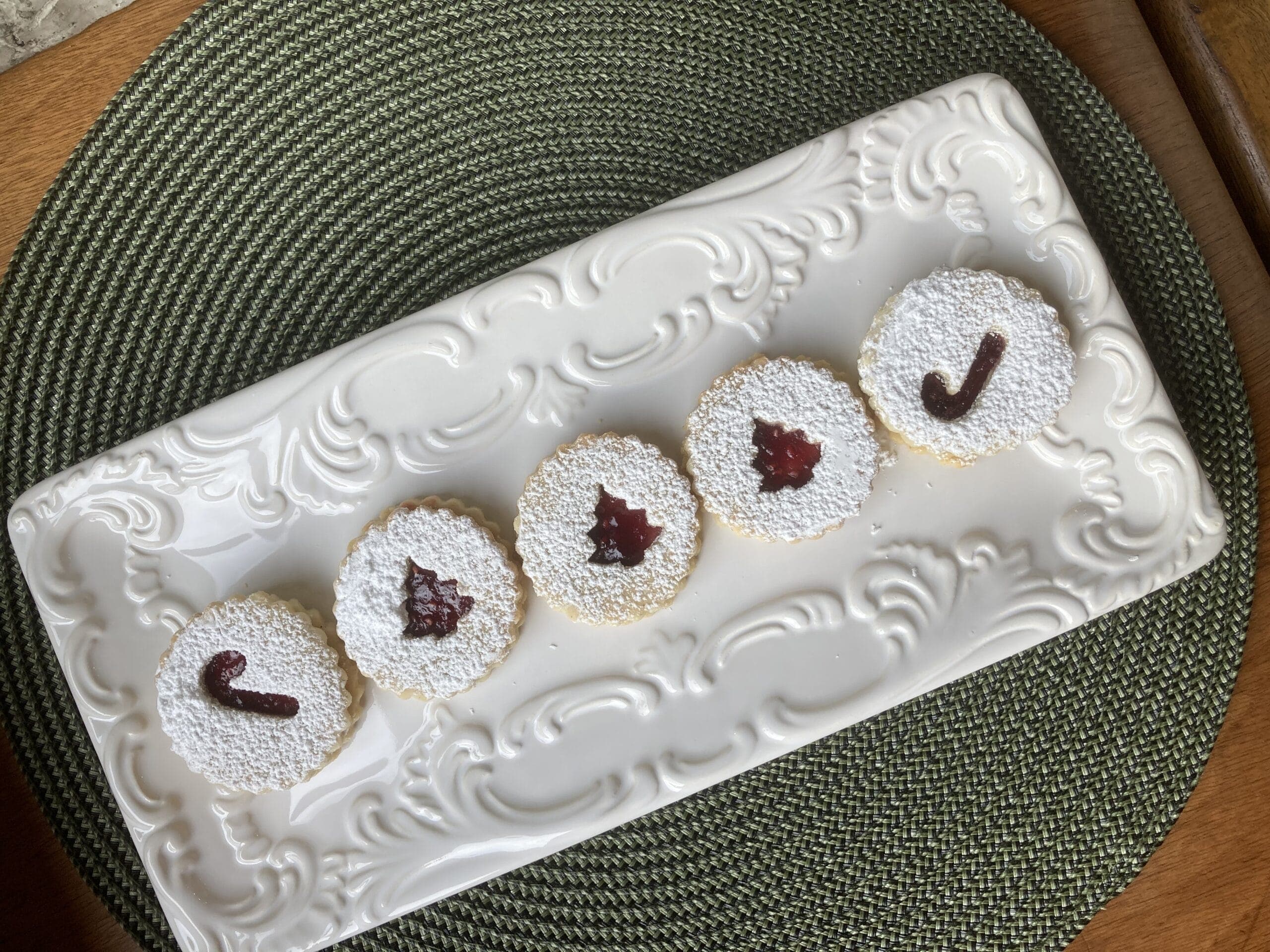 A decorative platter holds five round cookies dusted with powdered sugar and adorned with red fruit preserves in various shapes.