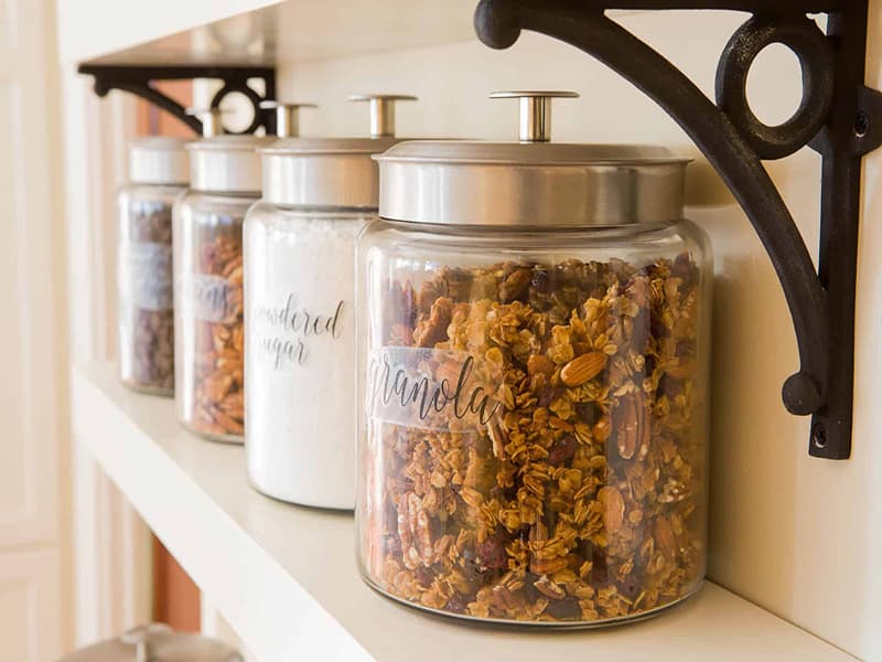 A shelf with glass jars labeled granola and powdered sugar, filled with assorted dry ingredients.
