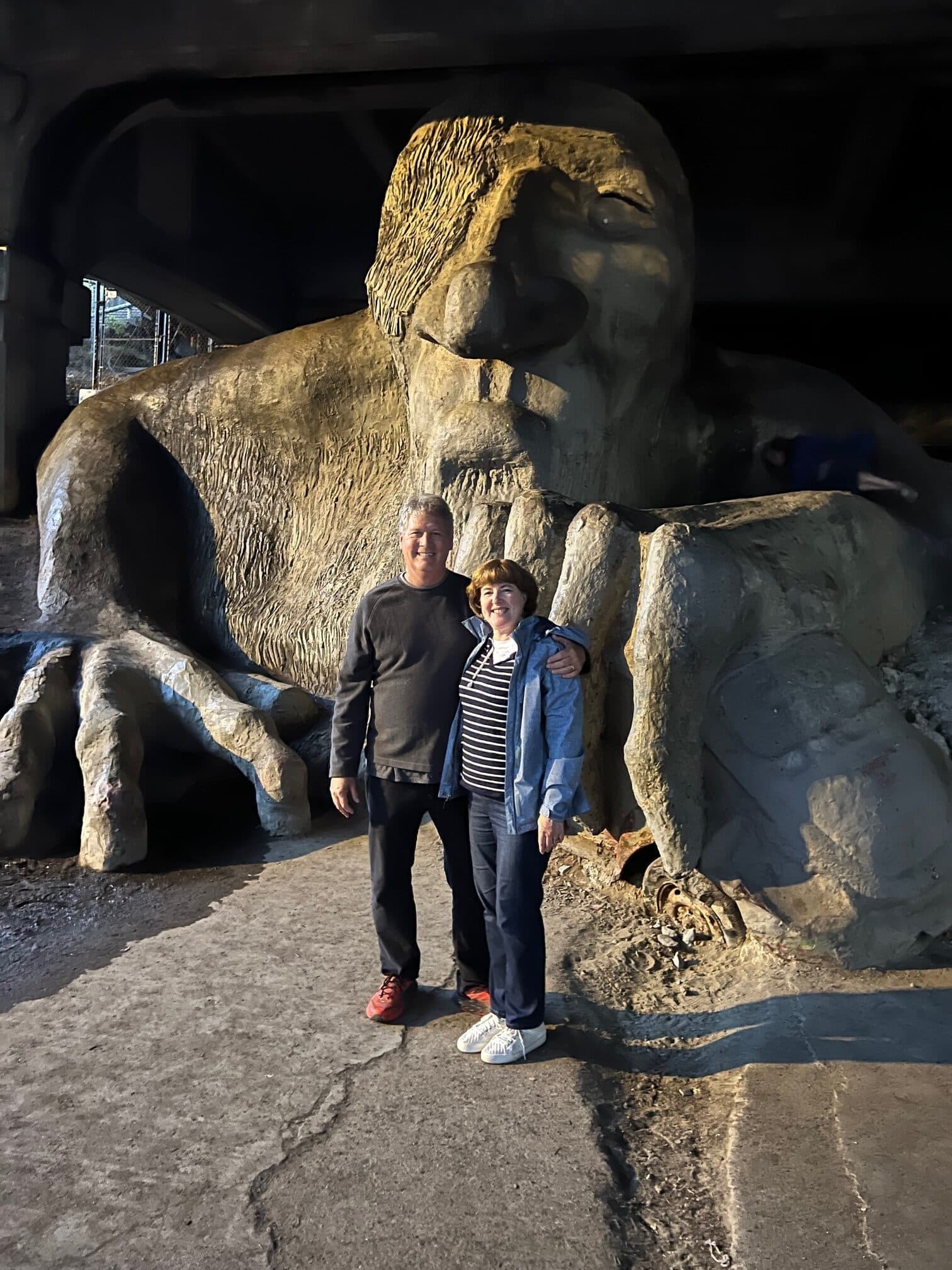 A man and a woman stand in front of a large concrete troll sculpture under a bridge.
