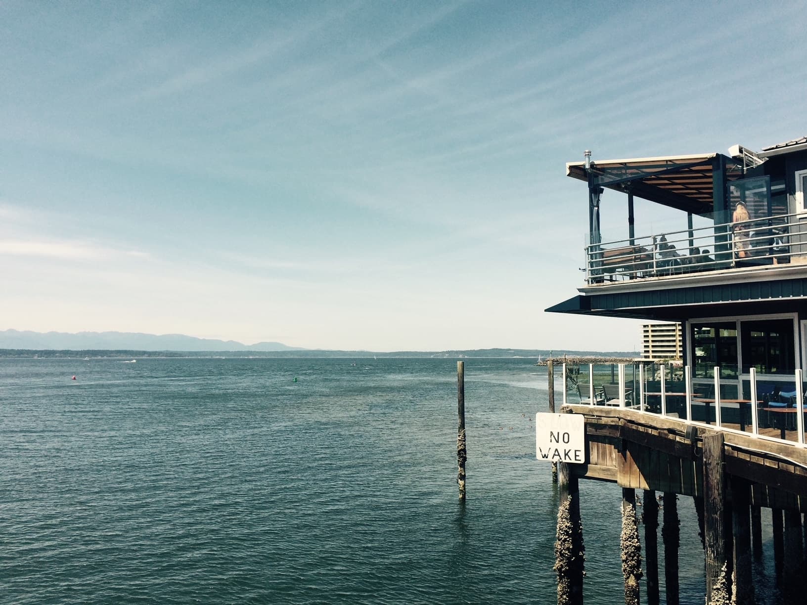 A wooden pier extends over calm waters with a "No Wake" sign, under a clear blue sky.