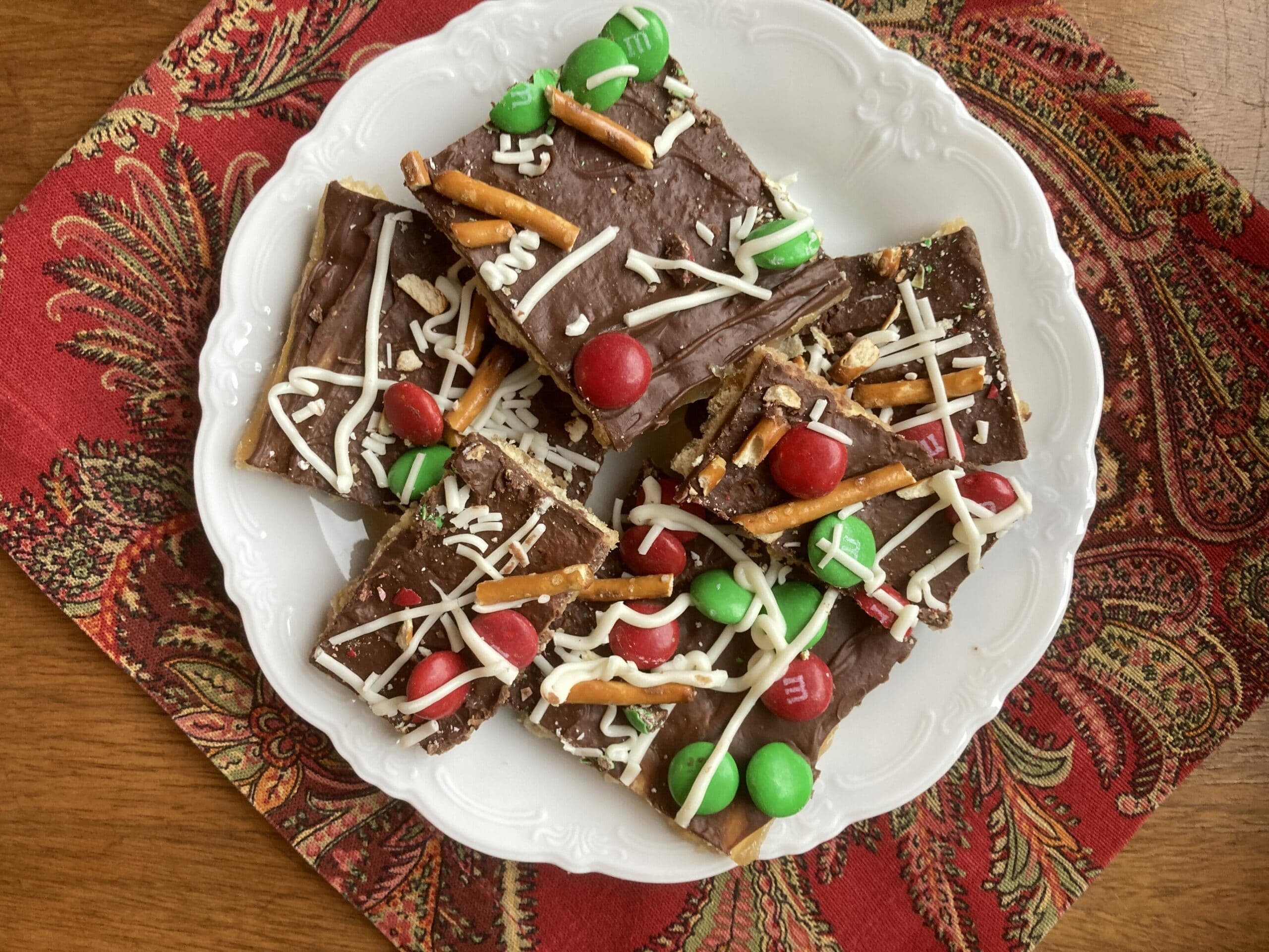 A plate of chocolate bark decorated with pretzels, red and green candies, and white drizzle, resting on a festive cloth.