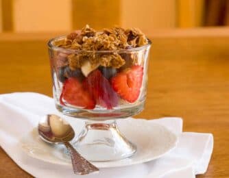 A glass dessert dish filled with layers of strawberries, blueberries, and granola, served with a spoon on a white napkin.