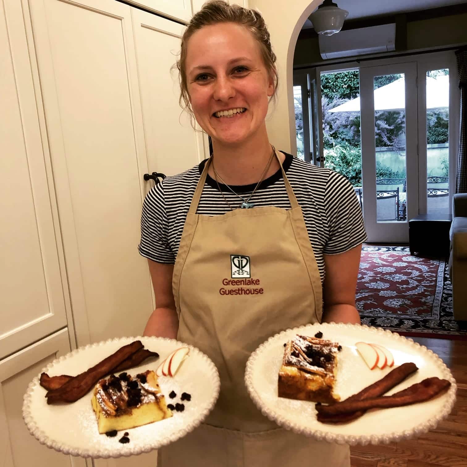 A smiling woman in an apron holds two plates of French toast and bacon in a cozy kitchen.