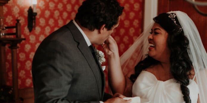 A newlywed couple shares a joyful moment, with the bride playfully touching the groom's face while both smile at each other.
