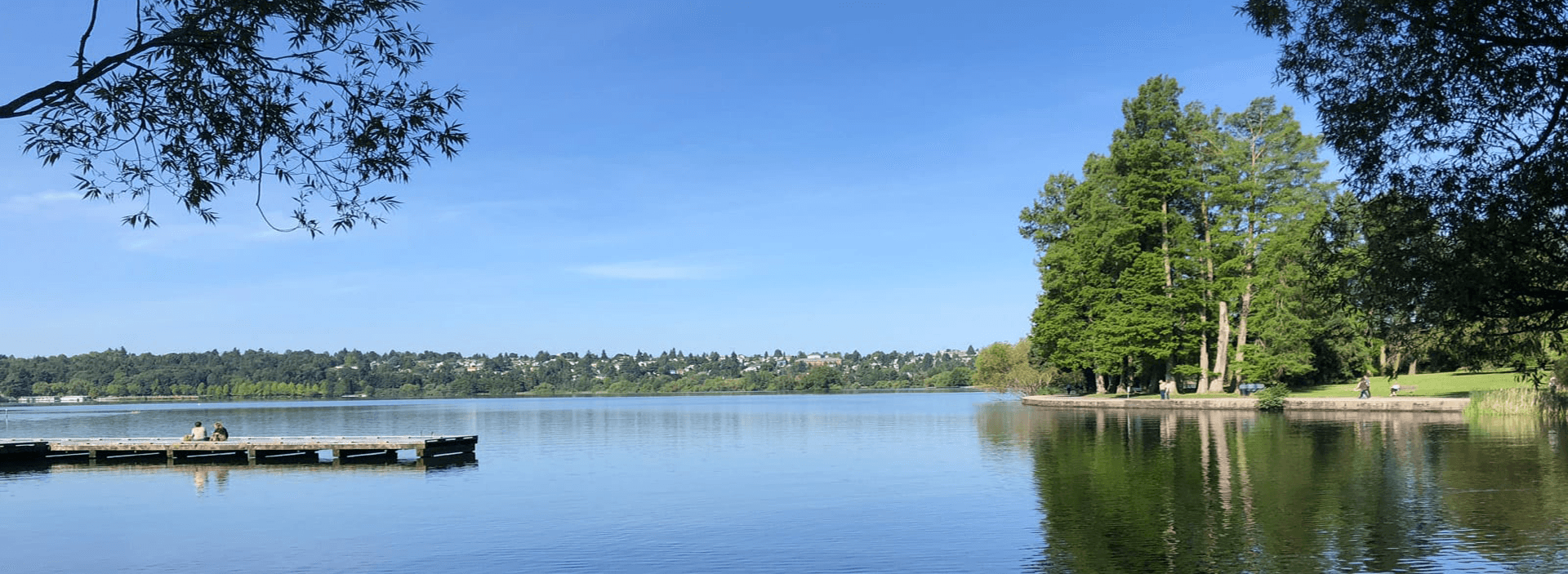 A peaceful lake scene with green trees, a wooden dock, and people walking along the shore under a clear blue sky.