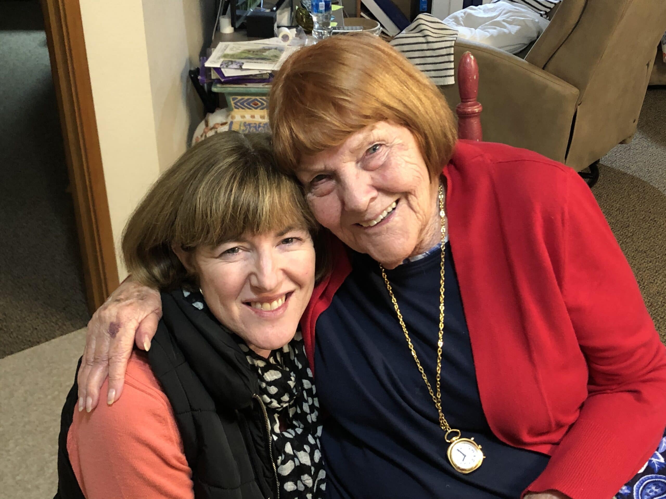 A woman sits closely with an elderly woman, both smiling warmly at the camera.