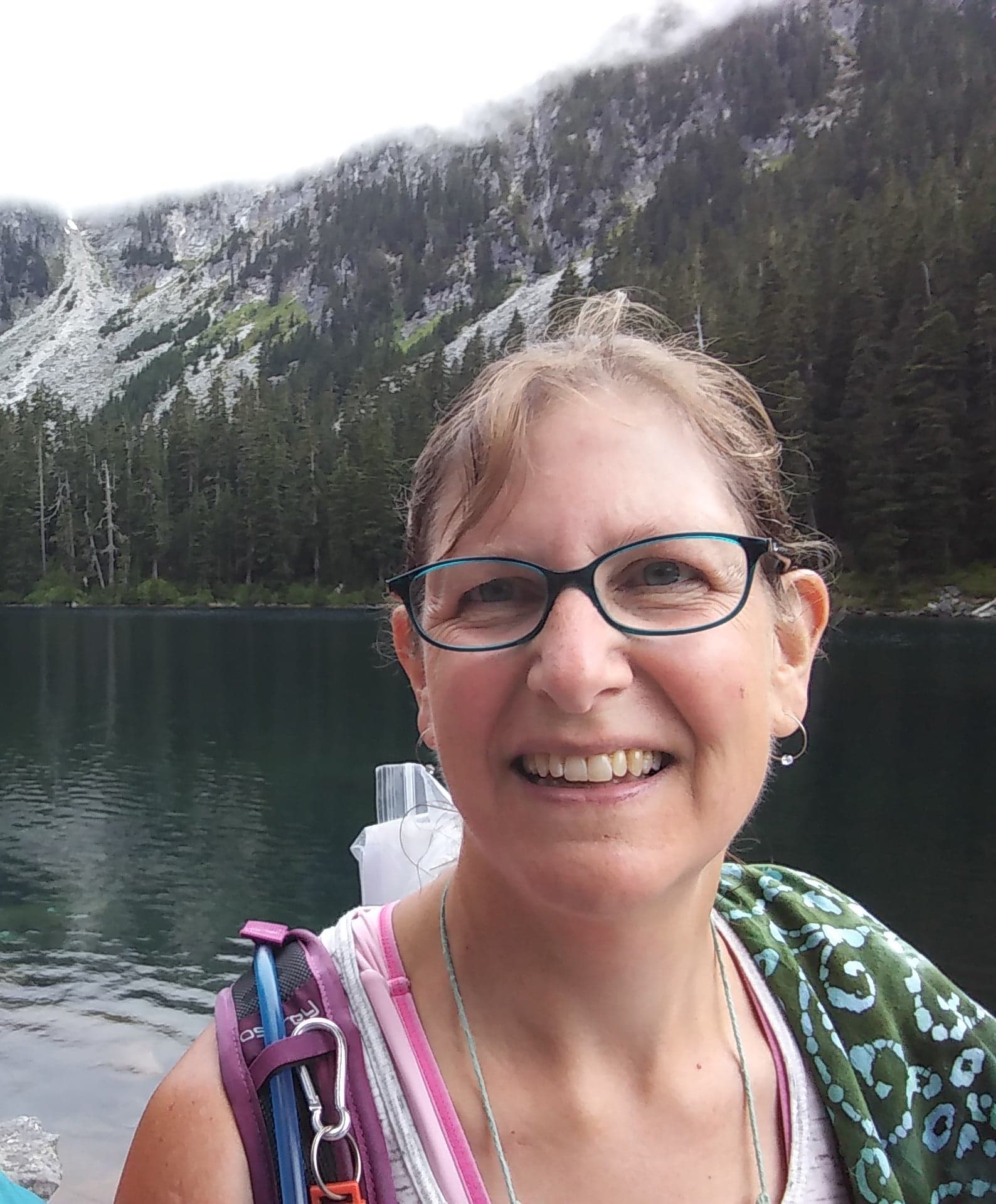 A smiling woman in glasses stands by a serene lake surrounded by mountains and trees.