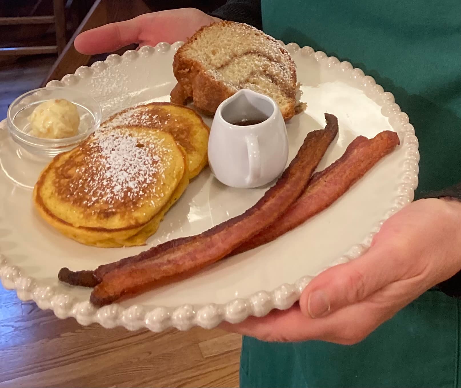 A person holds a plate with pancakes, a slice of bread, bacon, syrup, and butter.