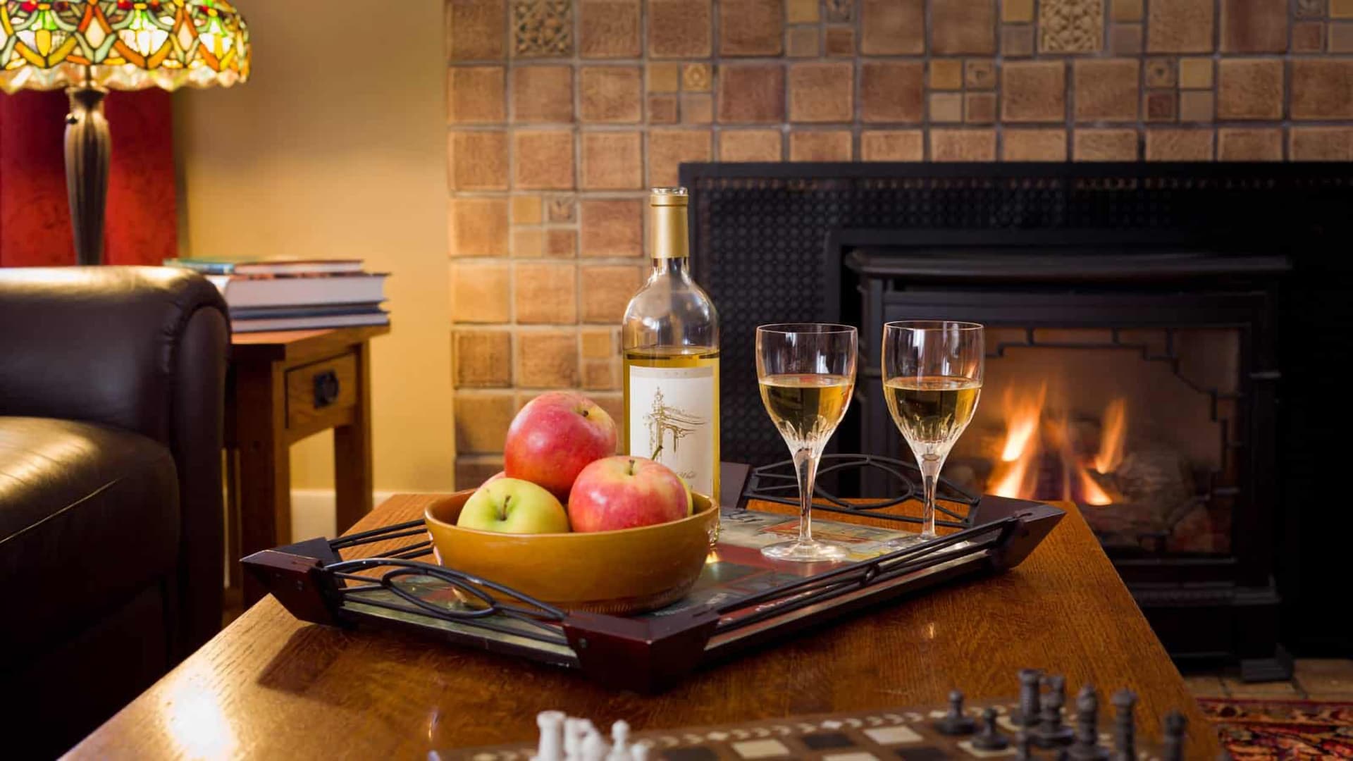 A cozy scene featuring two glasses of white wine, a bottle, apples in a bowl, and a warm fireplace.