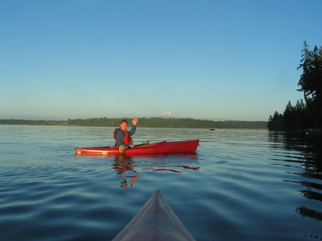 A person waves from a red kayak on a calm lake against a clear blue sky.