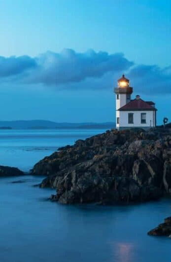 A lighthouse stands on rocky shore at twilight, with a calm sea and distant hills.