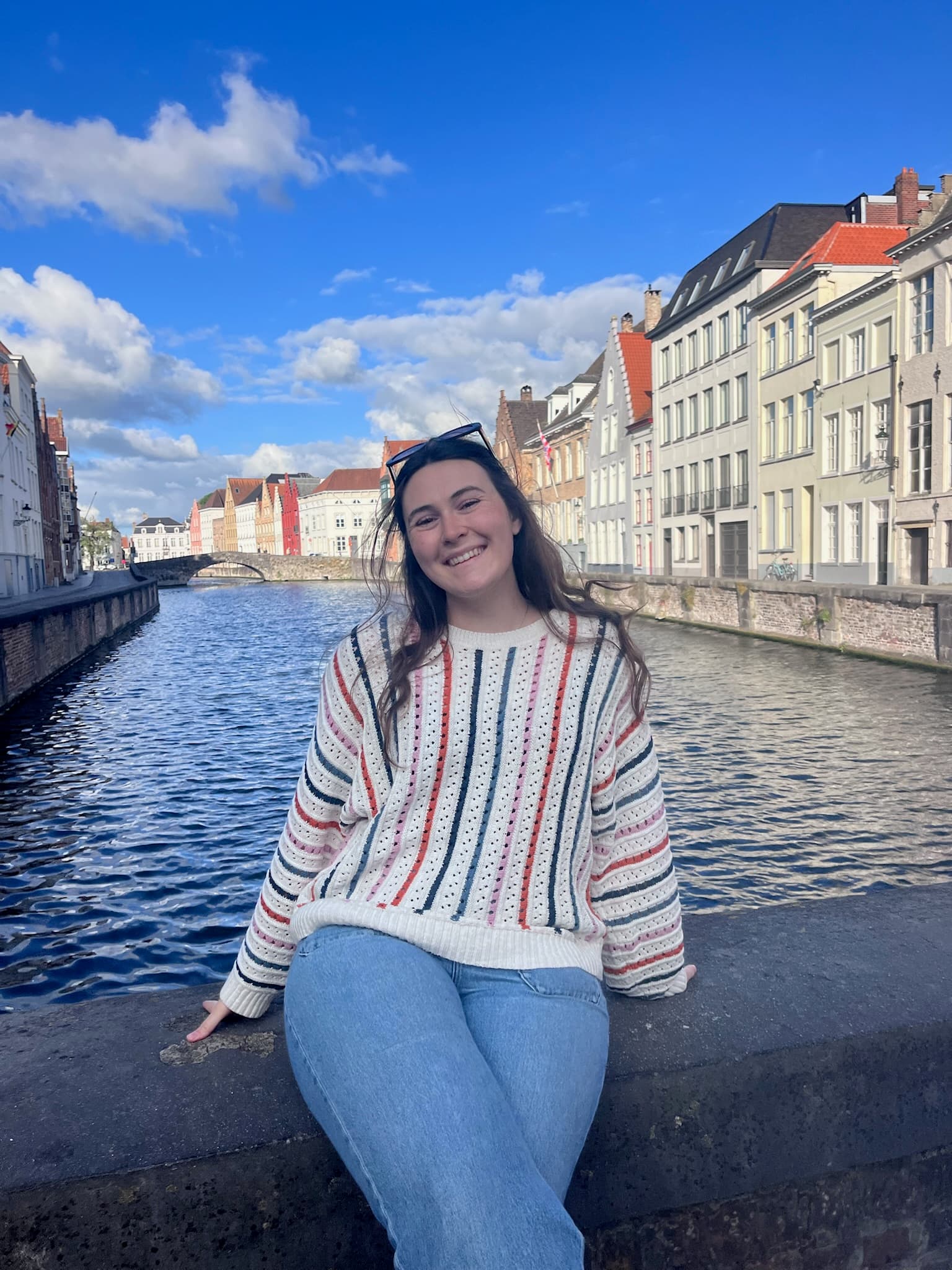 A woman smiles while sitting by a canal with colorful buildings in the background under a blue sky.