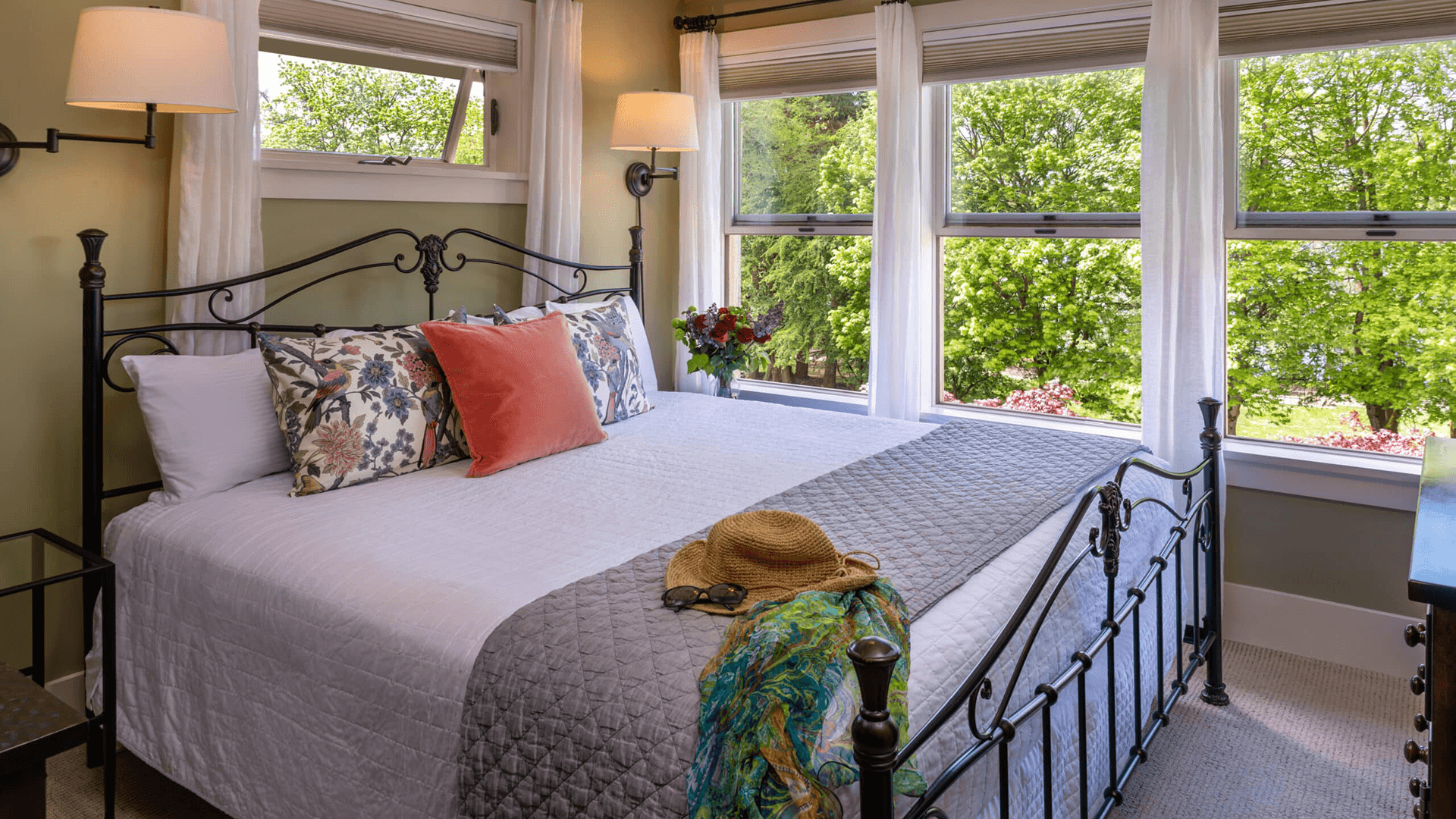 A cozy bedroom featuring a metal bed frame, decorative pillows, and large windows overlooking greenery.