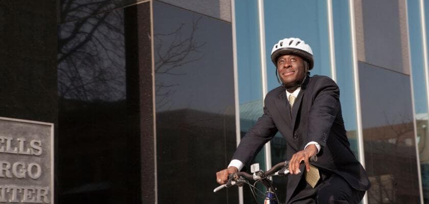 A man in a suit and helmet rides a bicycle near a modern glass building.