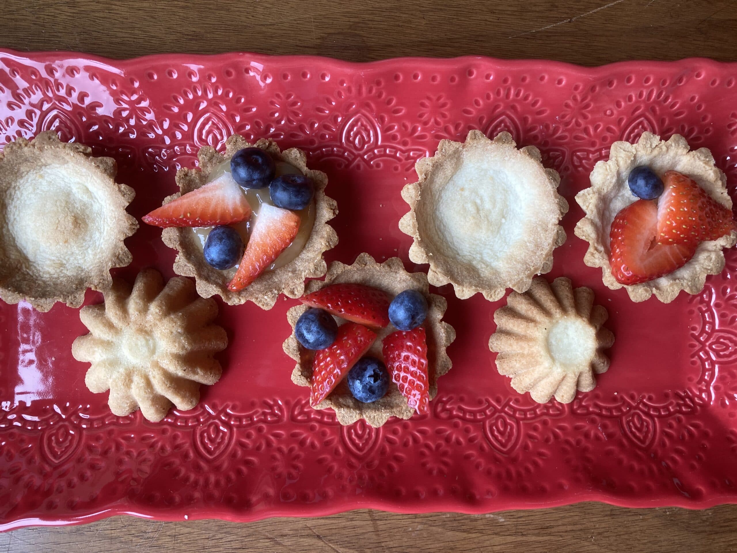 A red platter displaying various small tart shells topped with strawberries and blueberries.