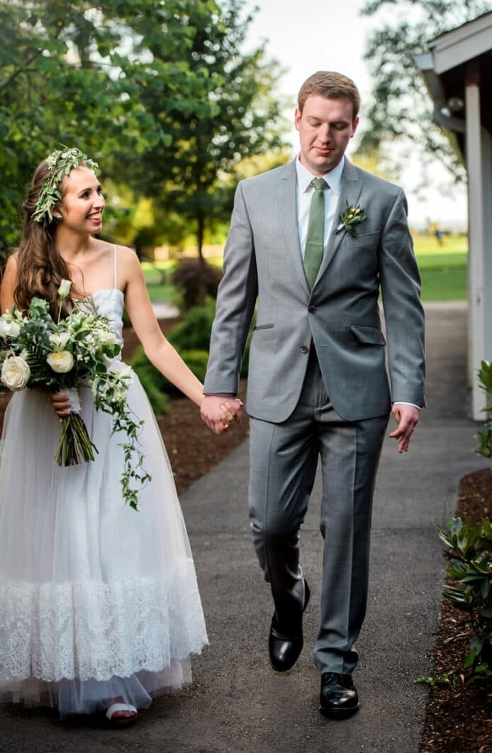 A smiling bride and groom hold hands while walking together on a path surrounded by greenery.