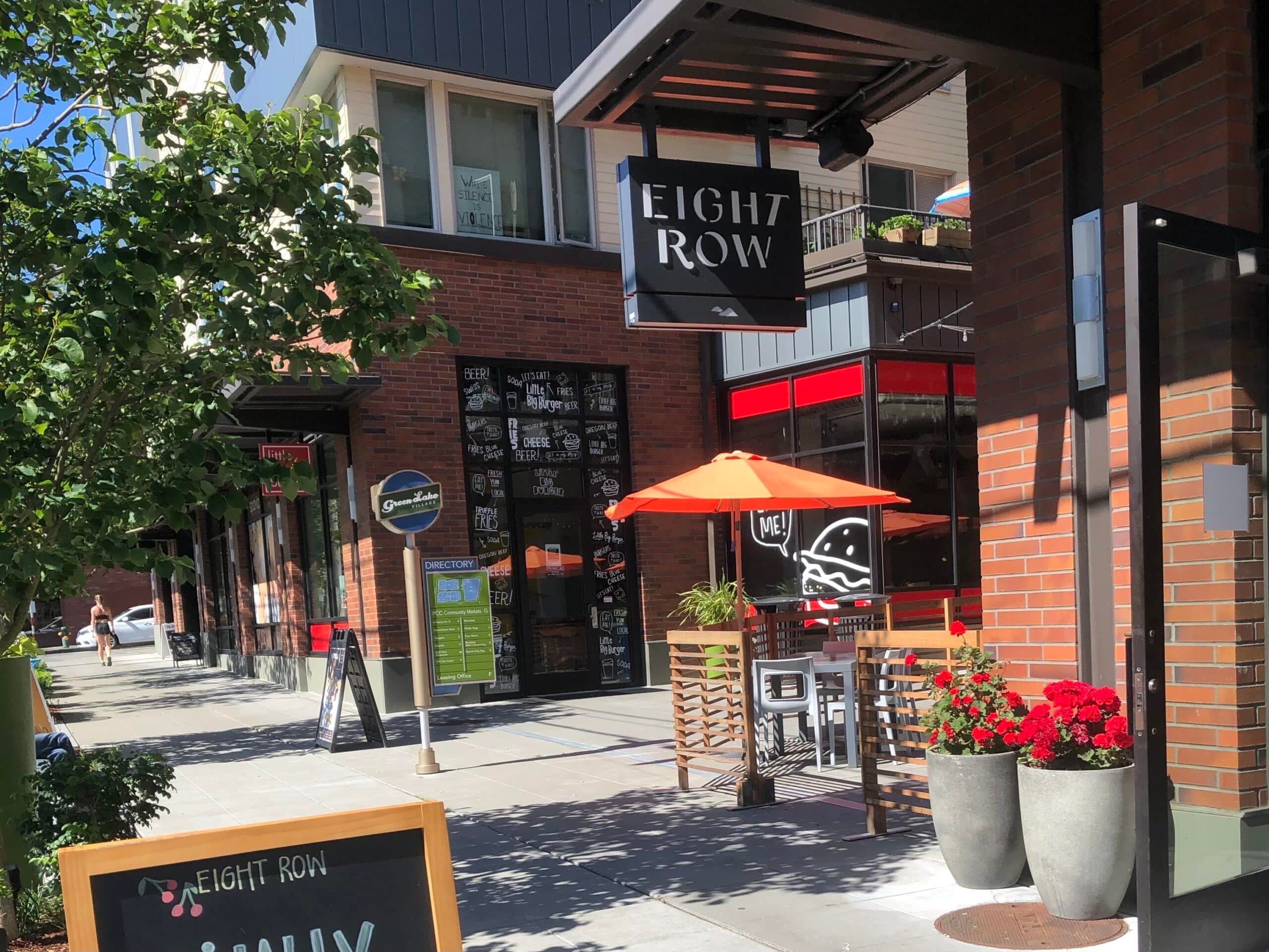A bright outdoor seating area in front of the restaurant "Eight Row" featuring red flowers and an orange umbrella.
