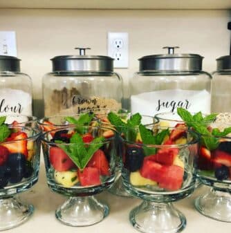 Three glass bowls filled with colorful fruit and mint leaves in front of labeled jars of flour, brown sugar, and sugar.