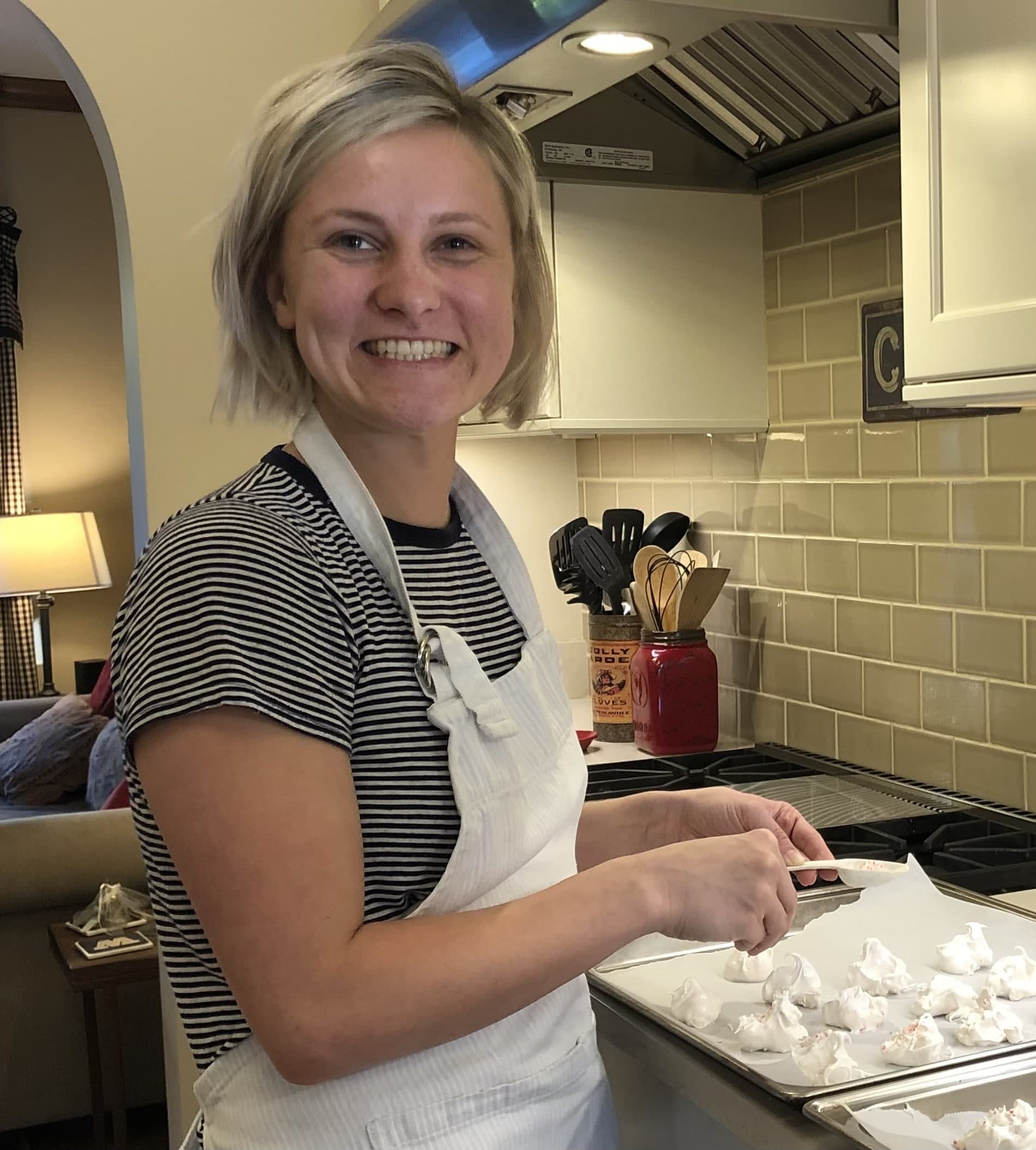 A smiling woman in a kitchen prepares meringue cookies on a baking sheet.