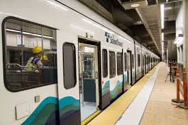 A subway train is stopped at a platform with a worker in a yellow hard hat visible inside.