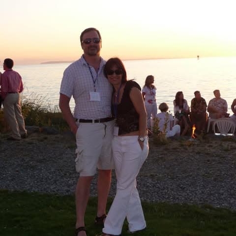 A couple stands together by the water at sunset, with others gathered in the background.