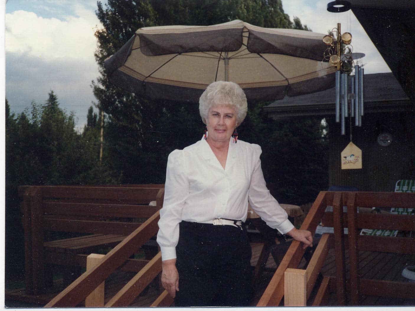 A woman with curly white hair stands on a wooden deck under a large umbrella, surrounded by trees.