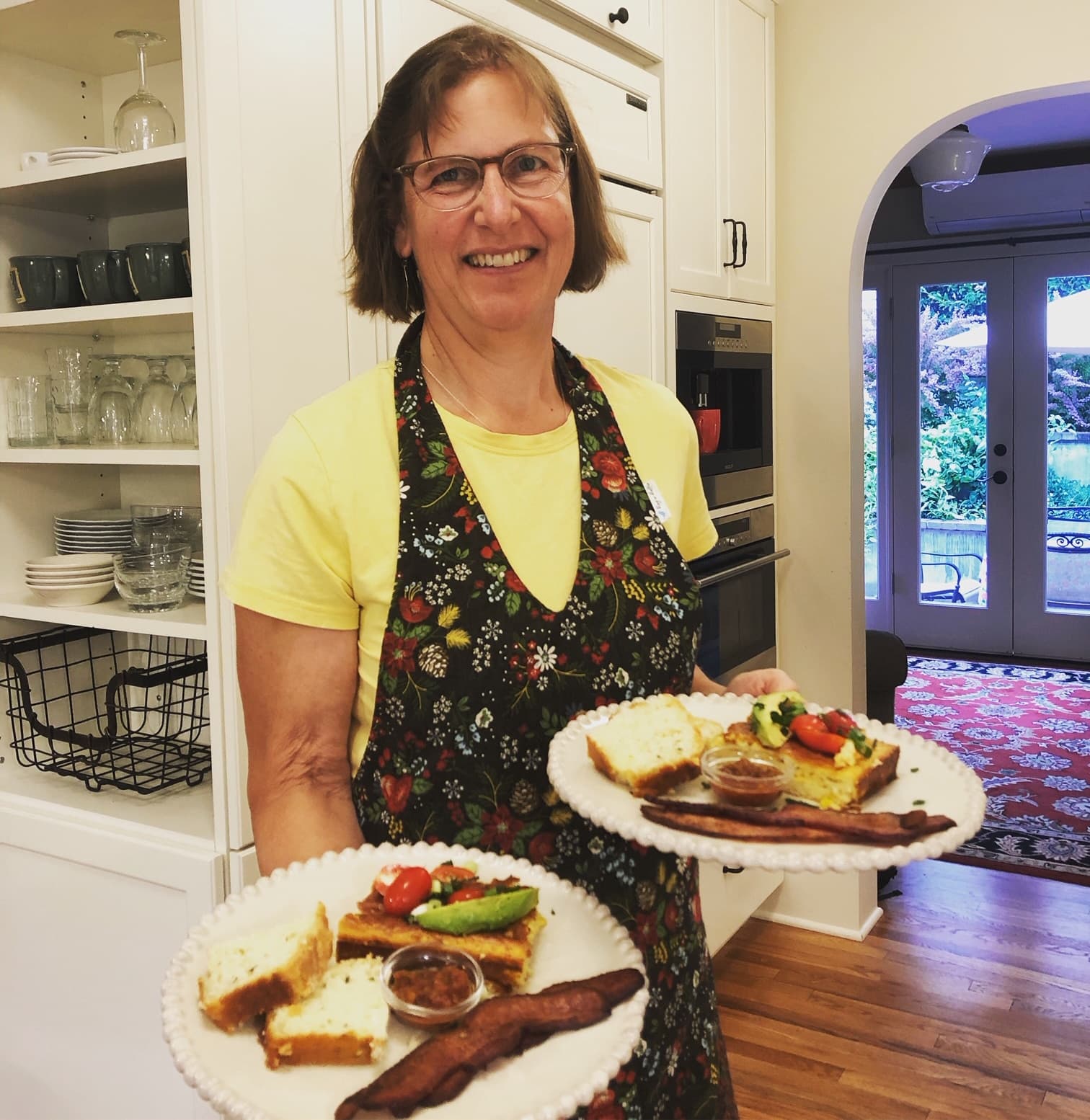 A woman in an apron smiles while holding two plates of food in a bright kitchen.