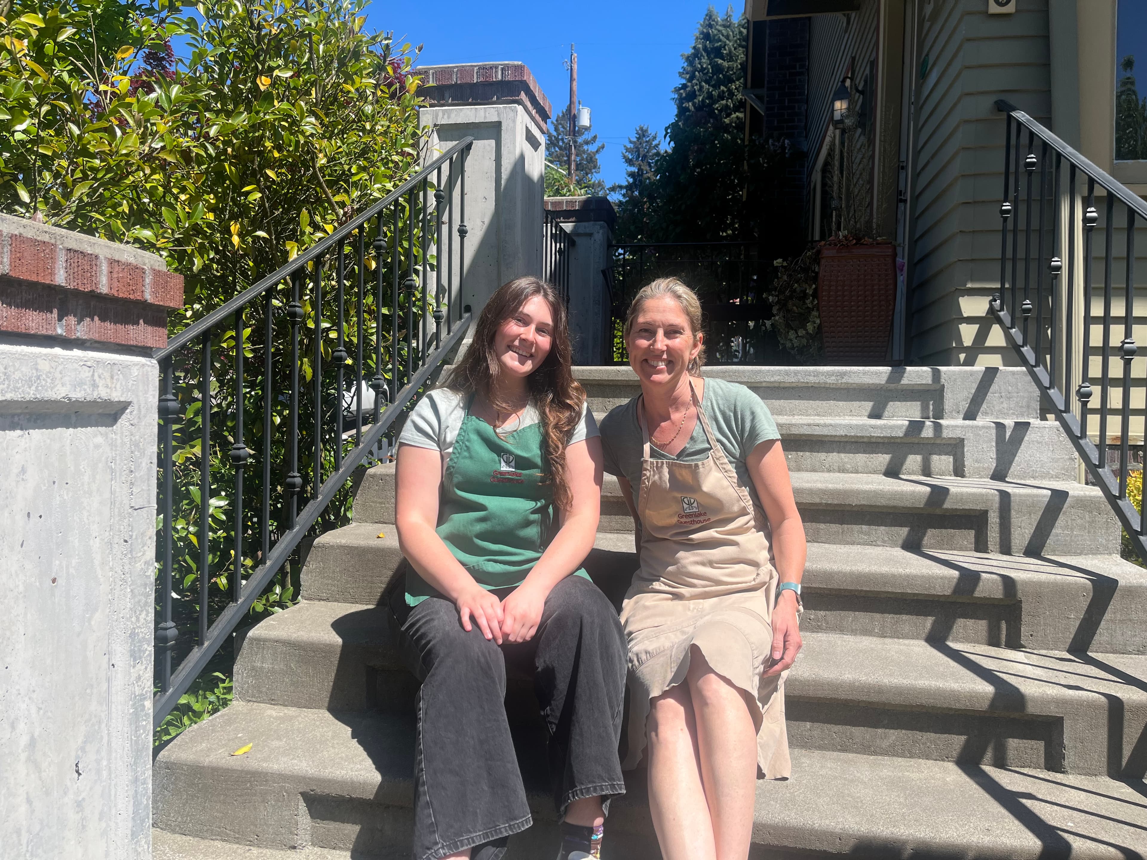 Two women in aprons sitting on outdoor steps, smiling in a sunny setting.