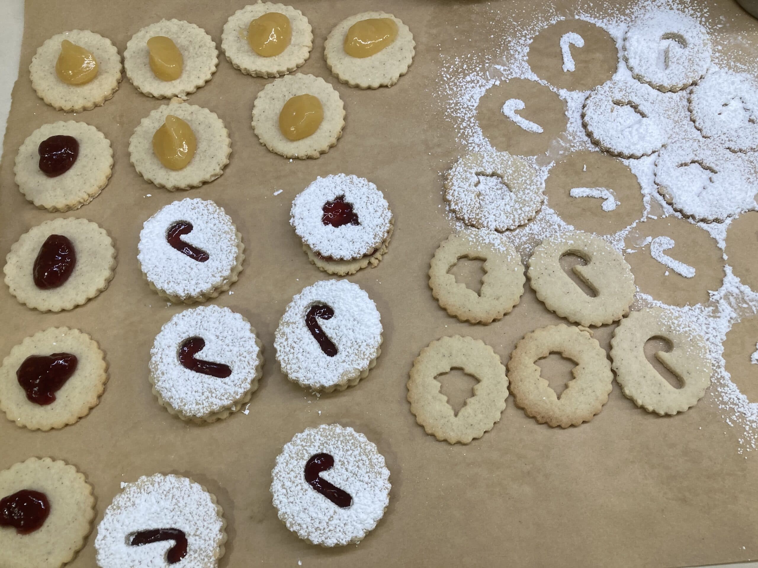 A variety of decorated cookies on a parchment-lined surface, featuring fruit preserves and powdered sugar designs.