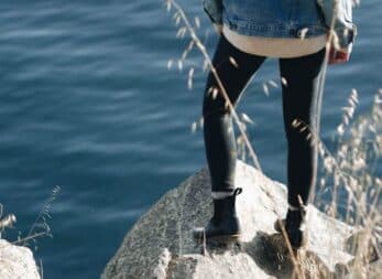A person stands on a rock by a body of water, wearing a denim jacket and black leggings.