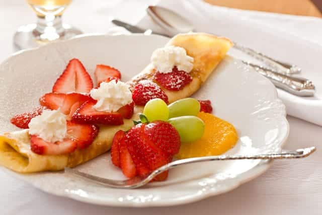 A plate of a strawberry-filled crepe garnished with whipped cream, alongside fresh fruit. A plate of a strawberry-filled crepe garnished with whipped cream, alongside fresh fruit.