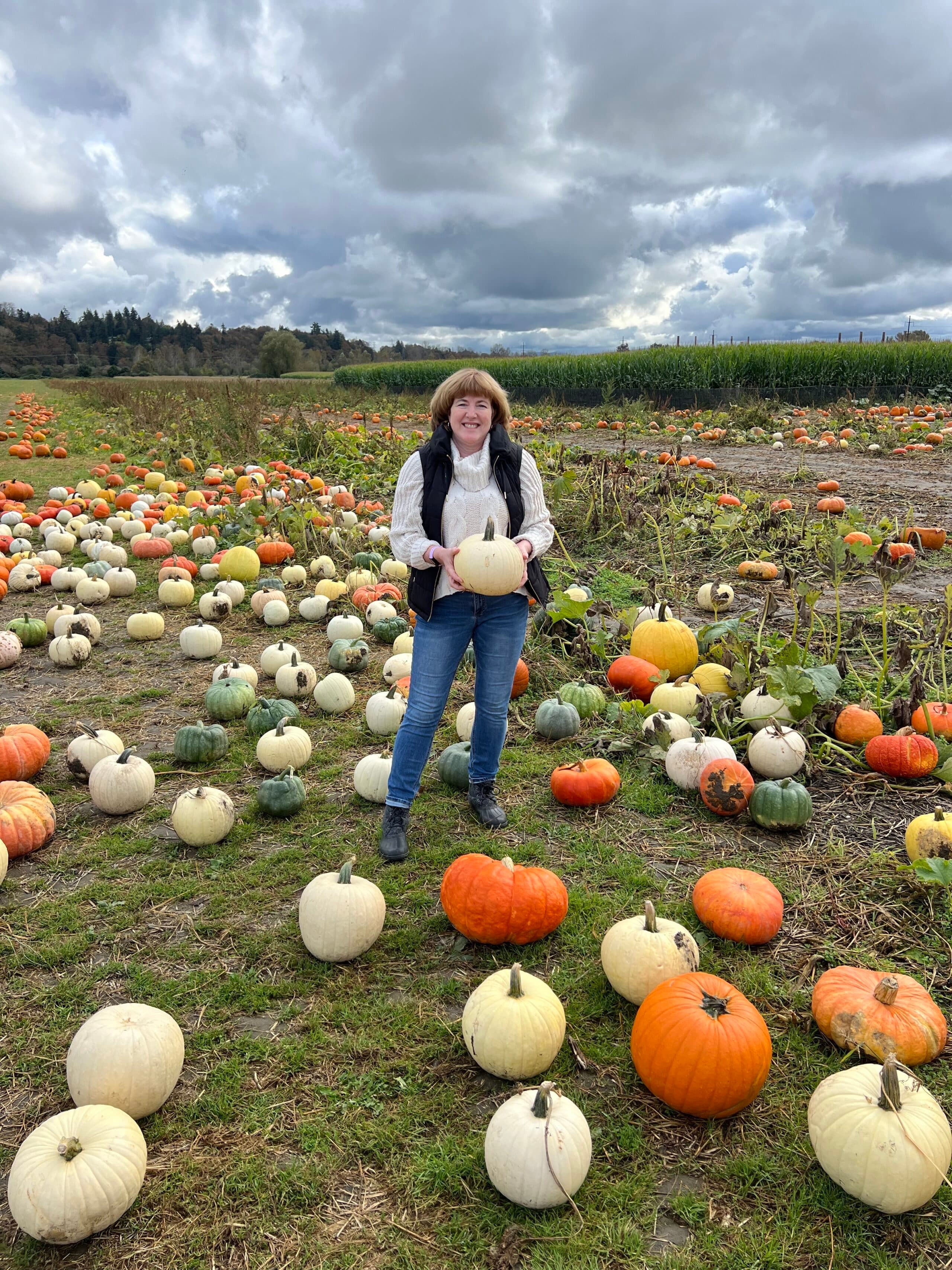 A woman stands in a pumpkin patch holding a white pumpkin, surrounded by various pumpkins under a cloudy sky.