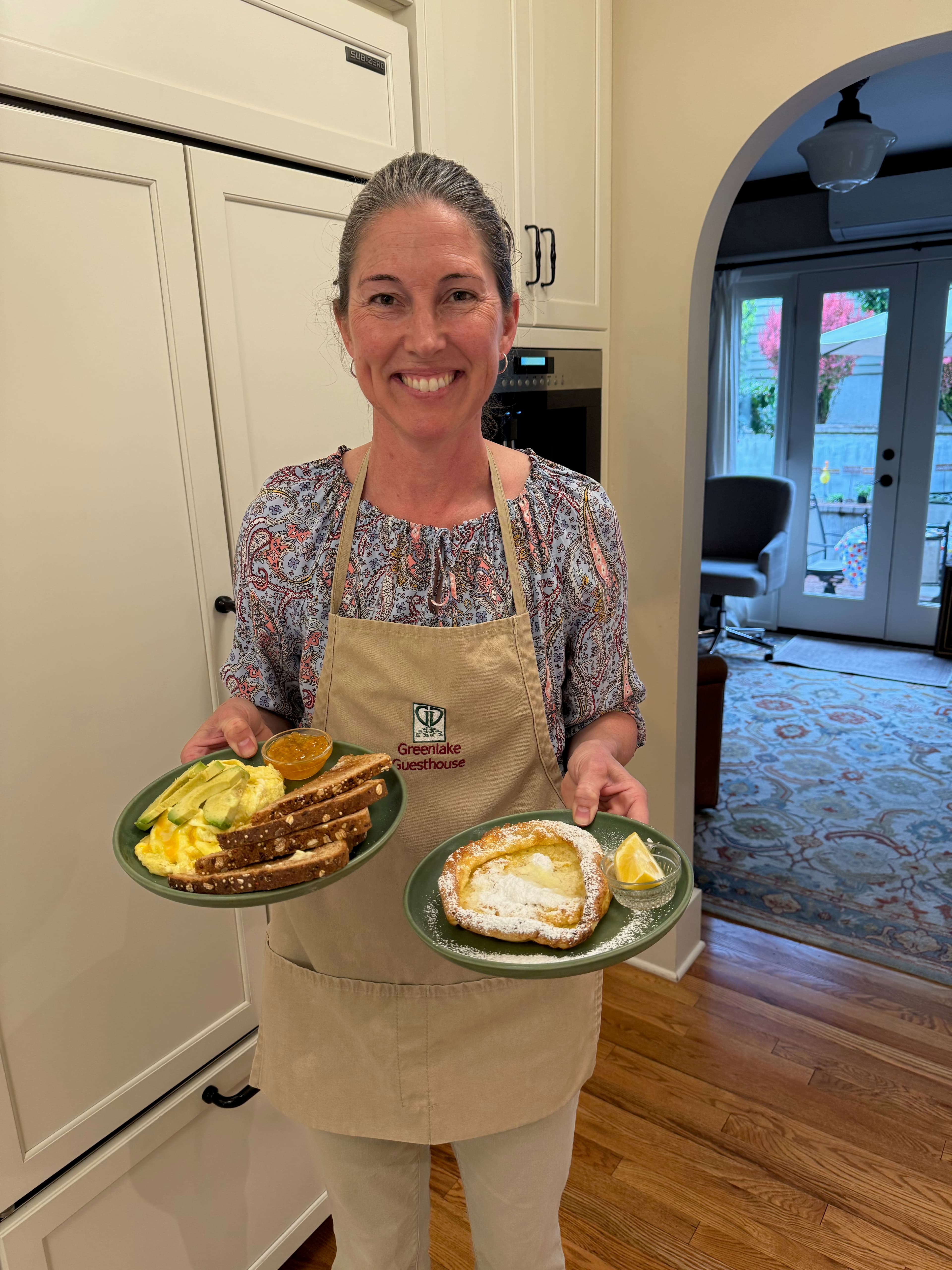 A smiling woman in an apron holds two plates of breakfast food in a kitchen.
