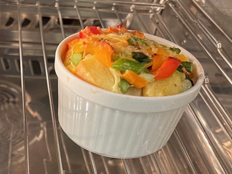 A white ramekin filled with baked vegetables sits inside an oven. A white ramekin filled with baked vegetables sits inside an oven.