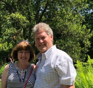 A man and woman smile together in a lush outdoor setting.