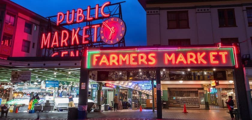 Neon signs for "PUBLIC MARKET" and "FARMERS MARKET" illuminate the entrance to a bustling market.