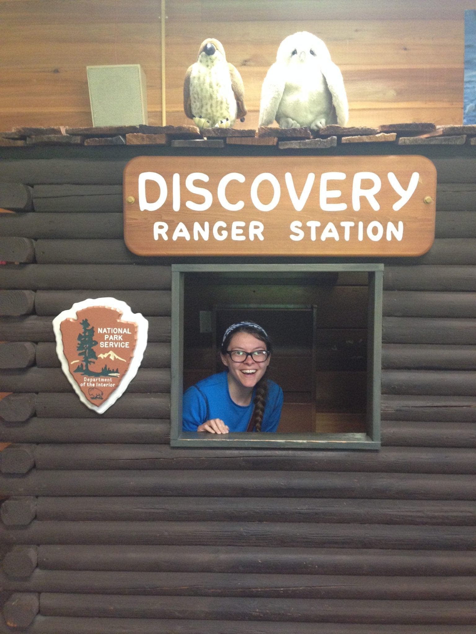 A ranger in a blue shirt smiles from the window of a Discovery Ranger Station, with two bird models overhead.