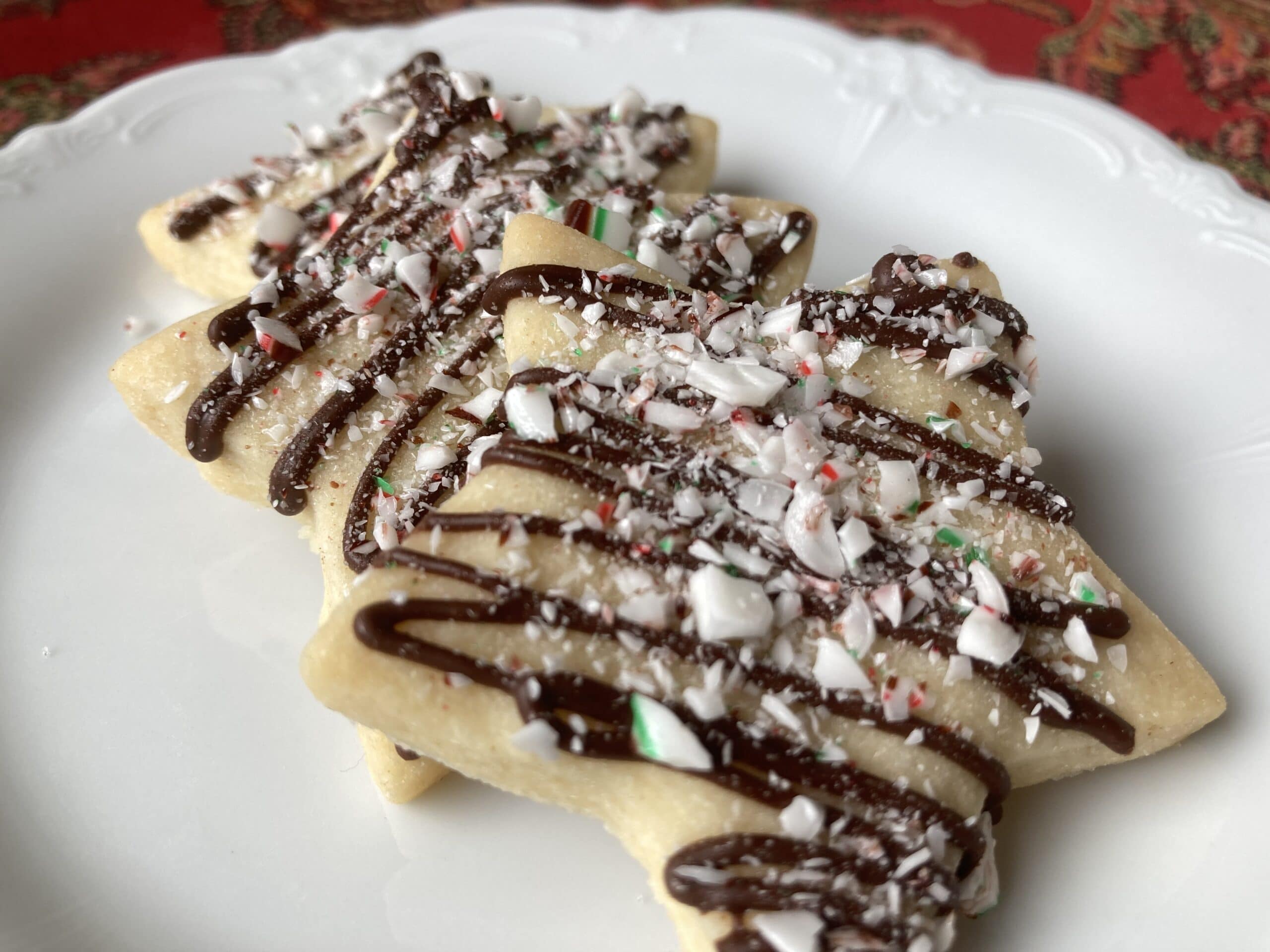 Three festive Christmas tree-shaped cookies drizzled with chocolate and topped with crushed peppermint on a decorative plate.